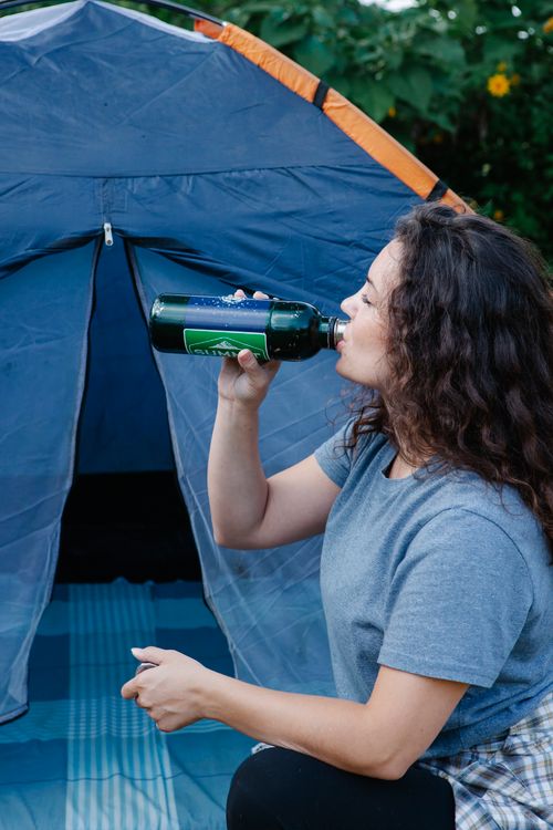 woman drinking water from bottle in front of blue tent at campsite
