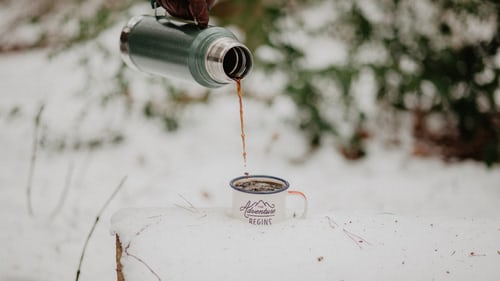 pouring hot coffee from insulated thermos into enamel mug on snow